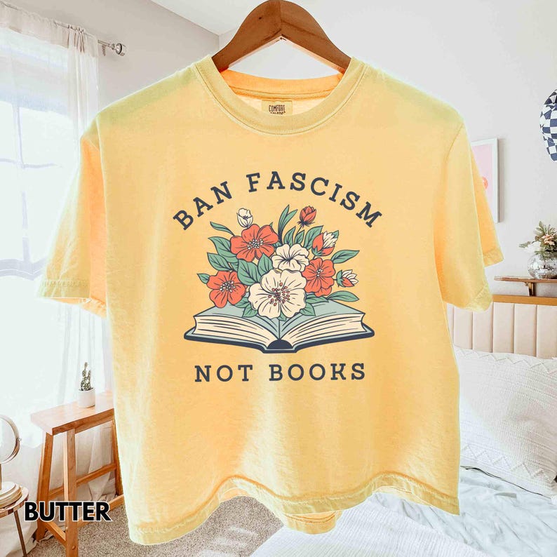 Op de afbeelding: Bottergeel t-shirt met de tekst "BAN FASCISM NOT BOOKS" boven een bloemenillustratie op een open boek. De bloemen zijn rood, wit en groen. Het shirt hangt aan een houten hanger.