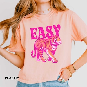 Peut inclure: T-shirt p&ecirc;che avec le mot "EASY" en lettres roses au-dessus d'une illustration de tigre rose et orange. Le mot "PEACHY" est imprim&eacute; en noir en bas &agrave; gauche du t-shirt.