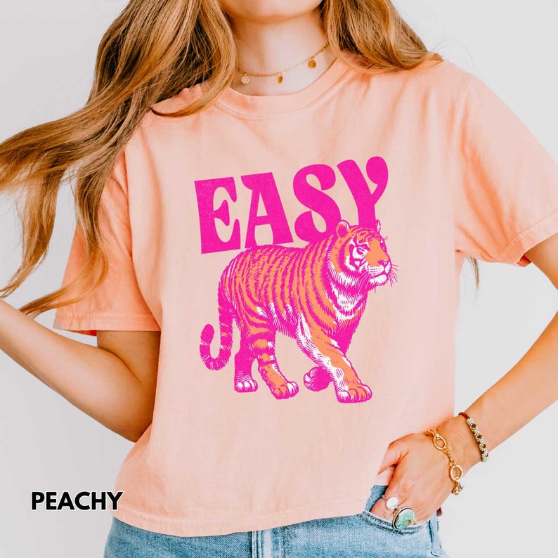 Peut inclure: T-shirt p&ecirc;che avec le mot "EASY" en lettres roses au-dessus d'une illustration de tigre rose et orange. Le mot "PEACHY" est imprim&eacute; en noir en bas &agrave; gauche du t-shirt.