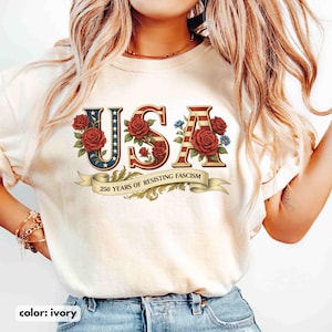 Op de afbeelding: Ivoor t-shirt met het woord "USA" in een patriottisch ontwerp, met rode rozen en blauwe bloemen. De "U" heeft een blauwe achtergrond met witte sterren, en de "S" en "A" hebben rood-witte strepen. Een banner leest "250 YEARS OF RESISTING FASCISM."