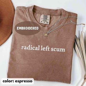 May include: Espresso-colored t-shirt with the phrase "radical left scum" in white lettering. The shirt has a crew neck and short sleeves. A silver necklace with a pendant rests on the shirt. The word "EMBROIDERED" is printed on the shirt.