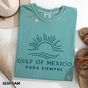 May include: Seafoam green t-shirt with a graphic of a sun setting over the ocean and the text "Gulf of Mexico Para Siempre".