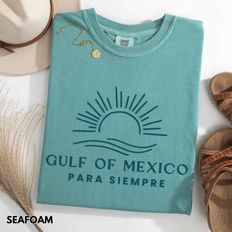 May include: Seafoam green t-shirt with a graphic of a sun setting over the ocean and the text "Gulf of Mexico Para Siempre".