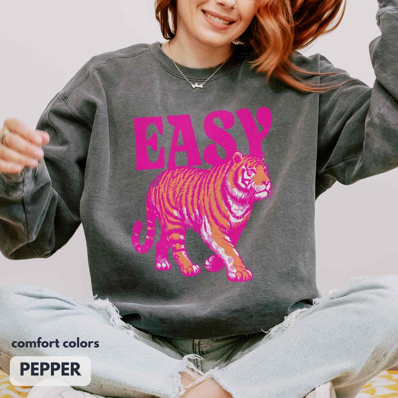 May include: A charcoal gray crewneck sweatshirt featuring the word EASY printed in bold pink letters above a graphic illustration of a tiger in shades of pink and orange. The garment has long sleeves and a ribbed neckline, cuffs, and hem.