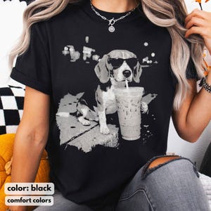 Puede incluir: Camiseta negra con un gr&aacute;fico en escala de grises de un beagle con gafas de sol y sosteniendo una bebida con pajita. El perro se representa en una pose relajada, lo que sugiere un estilo casual. Las palabras "color: black" y "comfort colors" est&aacute;n abajo.