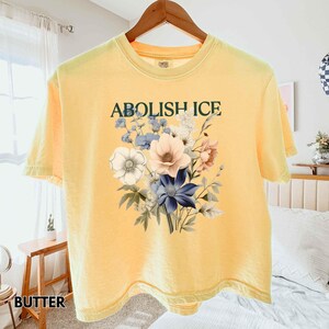 May include: Butter yellow t-shirt with the text "ABOLISH ICE" above a floral design. The floral arrangement includes white, pink, and blue flowers with green leaves. The shirt is hanging on a wooden hanger.