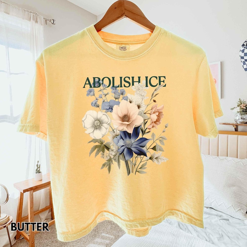May include: Butter yellow t-shirt with the text "ABOLISH ICE" above a floral design. The floral arrangement includes white, pink, and blue flowers with green leaves. The shirt is hanging on a wooden hanger.