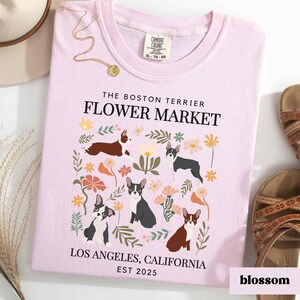 May include: Pink t-shirt with a floral design and Boston Terrier dogs. The text "The Boston Terrier Flower Market" is printed on the shirt, along with "Los Angeles, California Est 2025".