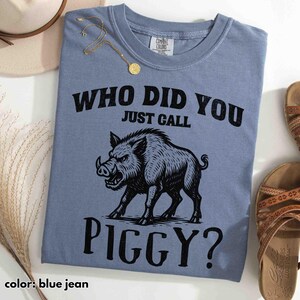 Op de afbeelding: Blauw spijkerbroekkleurig T-shirt met een zwarte afbeelding van een everzwijn en de tekst "WHO DID YOU JUST CALL PIGGY?". Het shirt is van het merk Comfort Colors. Een gouden ketting en sandalen zijn zichtbaar op de achtergrond.