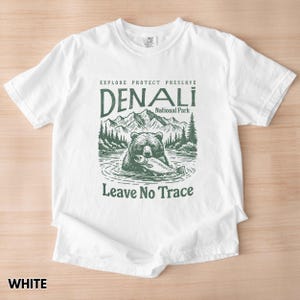 May include: White t-shirt with a green graphic of a bear fishing in a lake. The graphic includes the text "Explore Protect Preserve Denali National Park Leave No Trace".