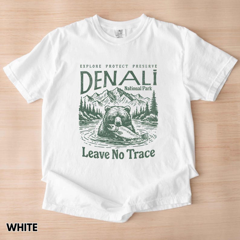 May include: White t-shirt with a green graphic of a bear fishing in a lake. The graphic includes the text "Explore Protect Preserve Denali National Park Leave No Trace".