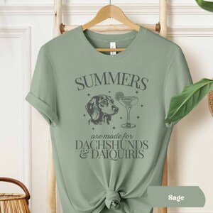 May include: Sage green t-shirt with a graphic of a dachshund and a cocktail glass. The text on the shirt reads "Summers are made for Dachshunds & Daiquiris".