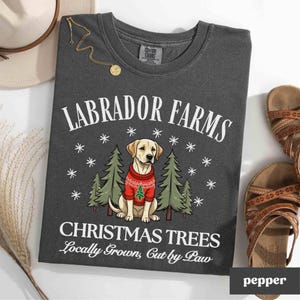 K&ouml;nnte beinhalten: Ein dunkelgraues T-Shirt mit dem Text "Labrador Farms Christmas Trees Locally Grown, Cut by Paw" und einem Cartoon-Bild eines gelben Labrador Retrievers, der einen roten Pullover mit einem kleinen Weihnachtsbaum tr&auml;gt.