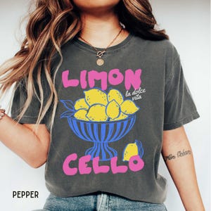May include: Dark gray t-shirt with a colorful graphic. The design features the word "LIMON" in pink above a blue bowl filled with yellow lemons. Below the bowl, the word "CELLO" is also in pink. The phrase "la dolce vita" is in white.