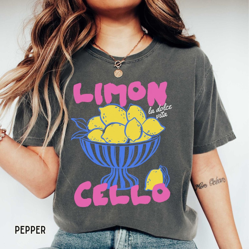 May include: Dark gray t-shirt with a colorful graphic. The design features the word "LIMON" in pink above a blue bowl filled with yellow lemons. Below the bowl, the word "CELLO" is also in pink. The phrase "la dolce vita" is in white.