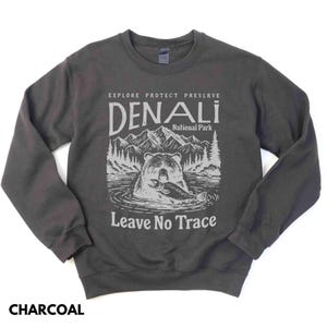 May include: A charcoal gray sweatshirt with a graphic design featuring a bear holding a fish in front of a mountain range. The text "Explore Protect Preserve Denali National Park Leave No Trace" is printed on the sweatshirt.