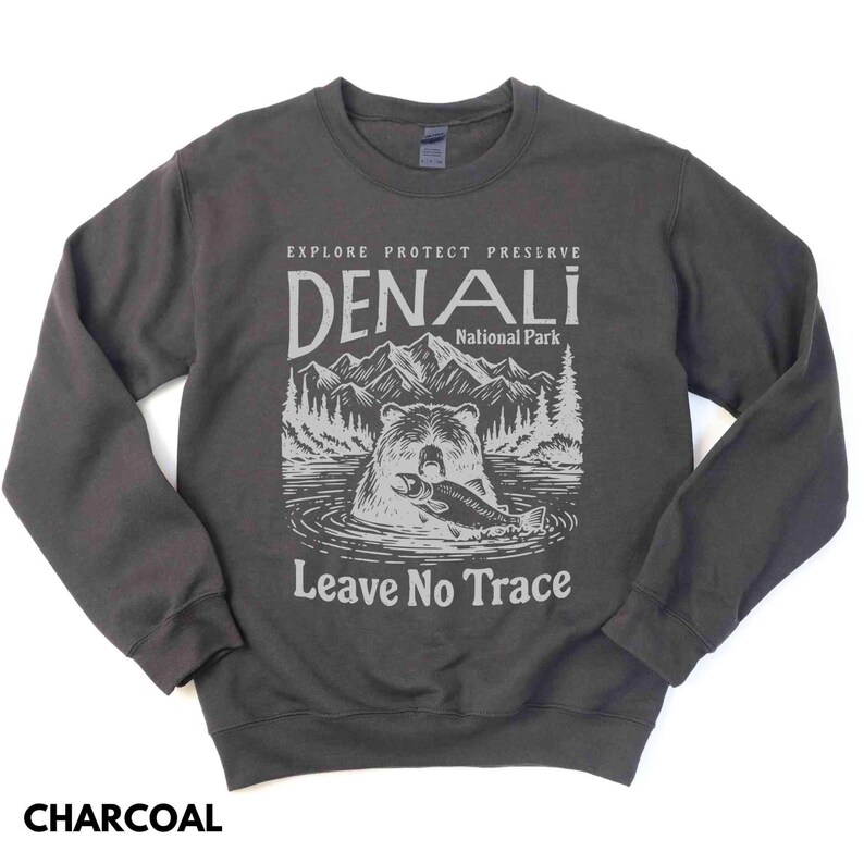 May include: A charcoal gray sweatshirt with a graphic design featuring a bear holding a fish in front of a mountain range. The text "Explore Protect Preserve Denali National Park Leave No Trace" is printed on the sweatshirt.