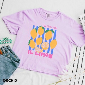 May include: Orchid-colored t-shirt with a lemon graphic. The design includes yellow lemons, blue stripes, and the text "La Dolce Vita" and "Il Limon". The shirt is paired with denim jeans and a checkered hair clip.