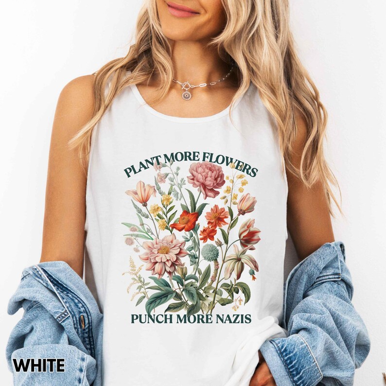 Op de afbeelding: Witte tanktop met een bloemenontwerp met roze, rode en gele bloemen. De tekst "PLANT MORE FLOWERS" en "PUNCH MORE NAZIS" is groen gedrukt. Het woord "WHITE" staat onderaan.