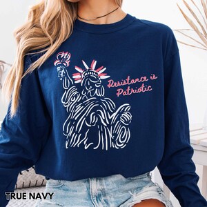 Pu&ograve; includere: Felpa a maniche lunghe blu navy con un disegno a tratto bianco della Statua della Libert&agrave;. Il testo "Resistance is Patriotic" &egrave; in rosa. La felpa &egrave; di colore blu navy.