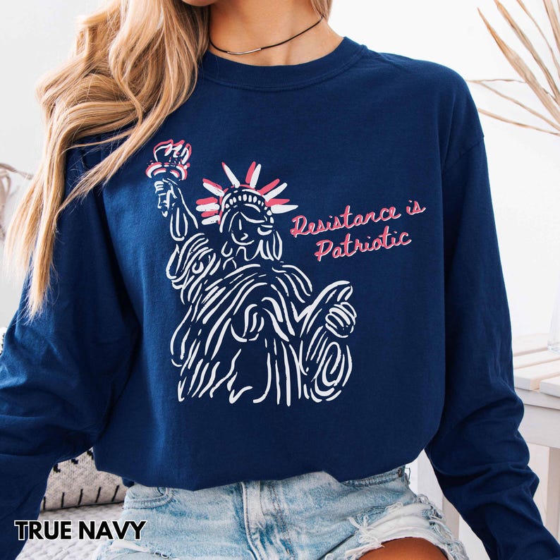Pu&ograve; includere: Felpa a maniche lunghe blu navy con un disegno a tratto bianco della Statua della Libert&agrave;. Il testo "Resistance is Patriotic" &egrave; in rosa. La felpa &egrave; di colore blu navy.