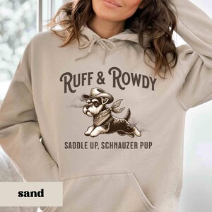 May include: A beige hoodie with a graphic of a dog wearing a cowboy hat and bandana. The text "Ruff & Rowdy Saddle Up, Schnauzer Pup" is printed on the hoodie.