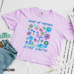 May include: A light purple t-shirt with a graphic print of a flamingo, flowers, and the text "Gulf of Mexico Stay Salty".
