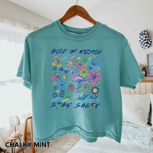 May include: A turquoise blue cropped t-shirt with a graphic print of a flamingo, flowers, and the text "Gulf of Mexico Stay Salty".