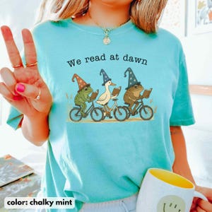 May include: Chalky mint-colored t-shirt with the text "We read at dawn." The shirt features a whimsical illustration of a frog, a duck, and a frog riding bicycles and reading books. The frogs and duck are wearing wizard hats.