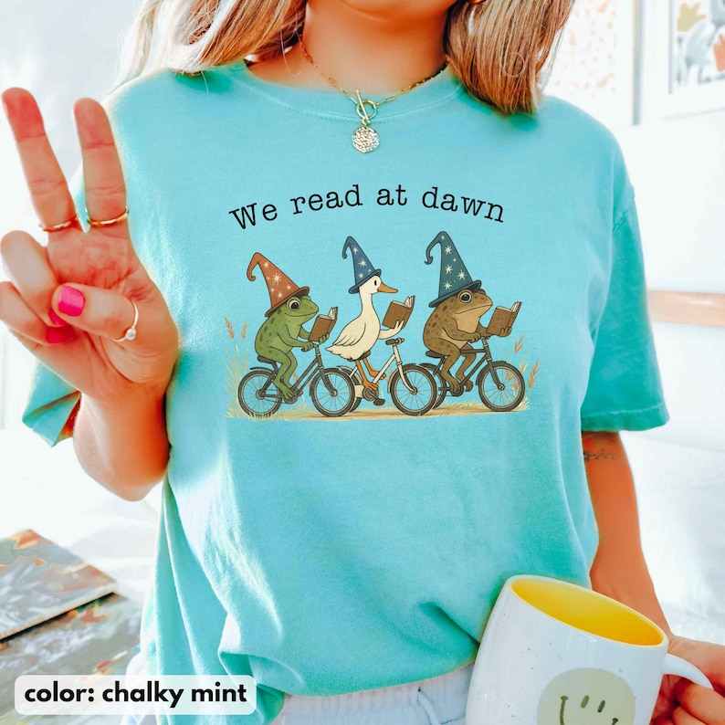 May include: Chalky mint-colored t-shirt with the text "We read at dawn." The shirt features a whimsical illustration of a frog, a duck, and a frog riding bicycles and reading books. The frogs and duck are wearing wizard hats.
