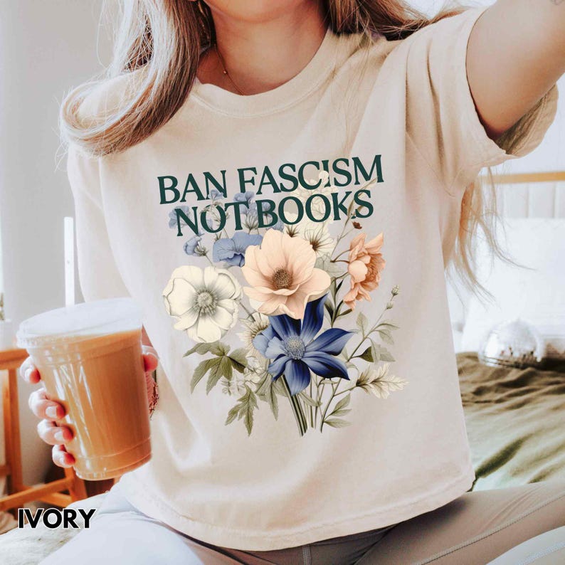 Puede incluir: Camiseta de color marfil con el texto "BAN FASCISM NOT BOOKS" en verde. La camiseta presenta un dise&ntilde;o floral con flores azules, melocot&oacute;n y blancas. La palabra "IVORY" est&aacute; impresa en la parte inferior.