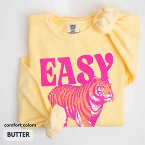 May include: A pale yellow crewneck sweatshirt featuring the word EASY in bold hot pink letters above a graphic of a tiger in shades of hot pink and orange. The inner neck label reads Comfort Colors with fabric content and origin details.