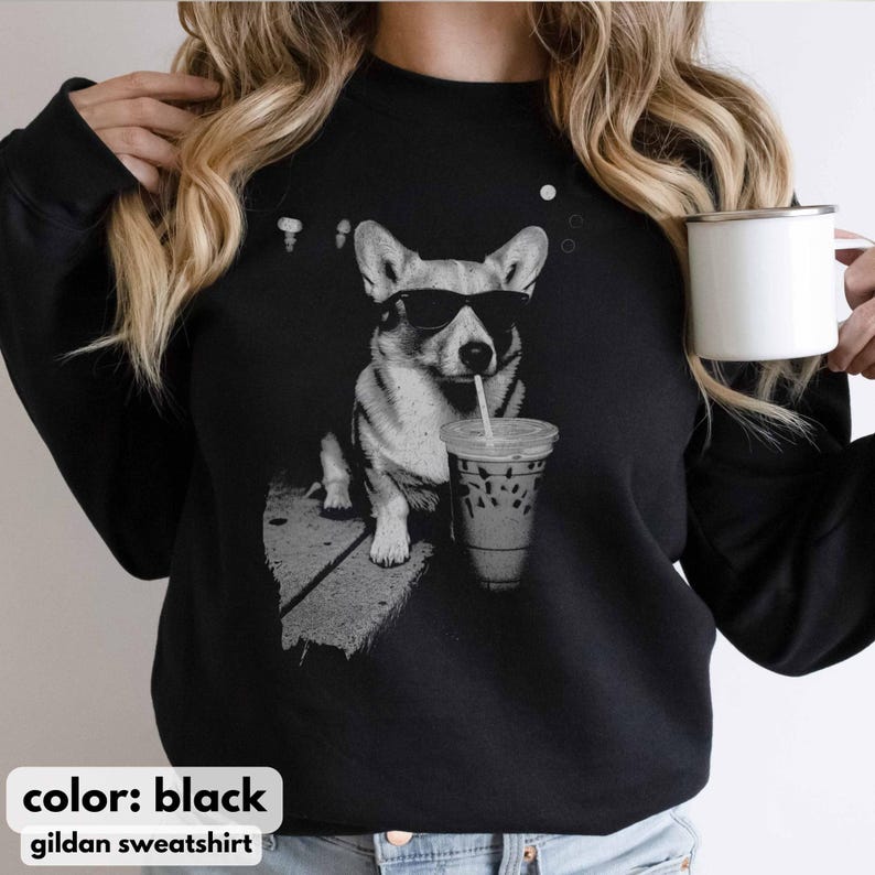 May include: Black Gildan sweatshirt featuring a grayscale graphic of a corgi wearing sunglasses and drinking from a beverage cup with a straw. The text "color: black" and "gildan sweatshirt" are at the bottom.
