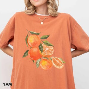 以下が含まれることがあります： オレンジ色のTシャツに、オレンジのグラフィックと、各オレンジに「Fruit Tramp」という文字が繰り返されて描かれています。