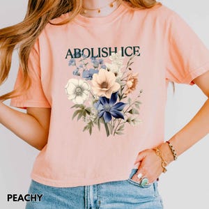 May include: Peach-colored t-shirt with the text "ABOLISH ICE" above a floral bouquet. The bouquet features white, blue, and peach-colored flowers. The word "PEACHY" is printed at the bottom of the shirt.