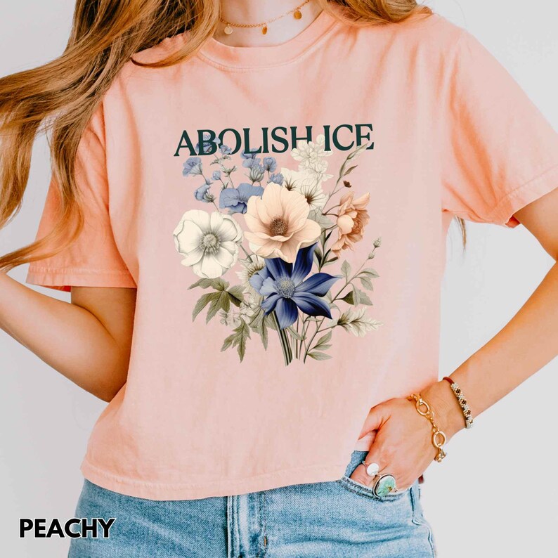 May include: Peach-colored t-shirt with the text "ABOLISH ICE" above a floral bouquet. The bouquet features white, blue, and peach-colored flowers. The word "PEACHY" is printed at the bottom of the shirt.