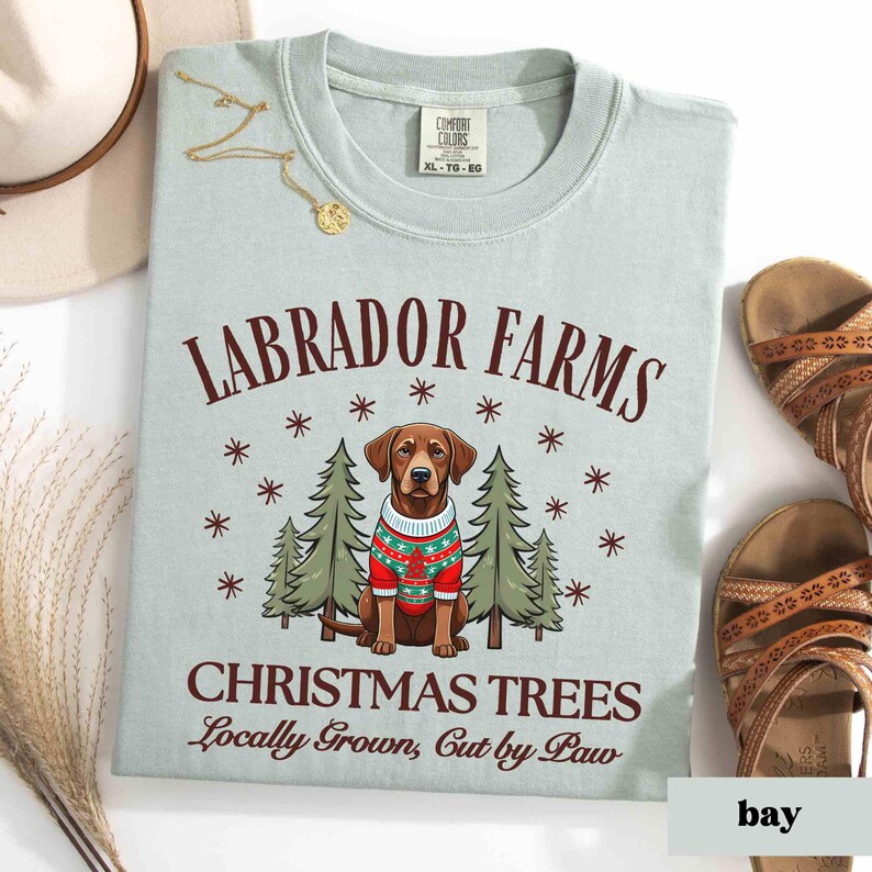 K&ouml;nnte beinhalten: Ein hellblaues T-Shirt mit dem Text "Labrador Farms Christmas Trees Locally Grown, Cut by Paw" in brauner Schrift. Ein Cartoon-Labrador Retriever, der einen rot-wei&szlig;en Pullover tr&auml;gt, steht vor zwei gr&uuml;nen Weihnachtsb&auml;umen.