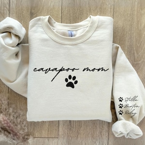 May include: A cream-colored sweatshirt with the text "cavapoo mom" and a paw print. The sleeve has three paw prints with the names "Stella", "Benton", and "Riley" written underneath.