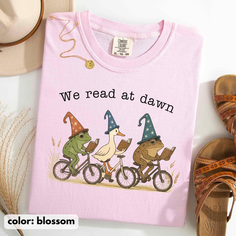 May include: Blossom pink Comfort Colors sweatshirt with the text "We read at dawn." The graphic features a frog, a duck, and another frog riding bicycles and reading books. A gold necklace and sandals are also visible.