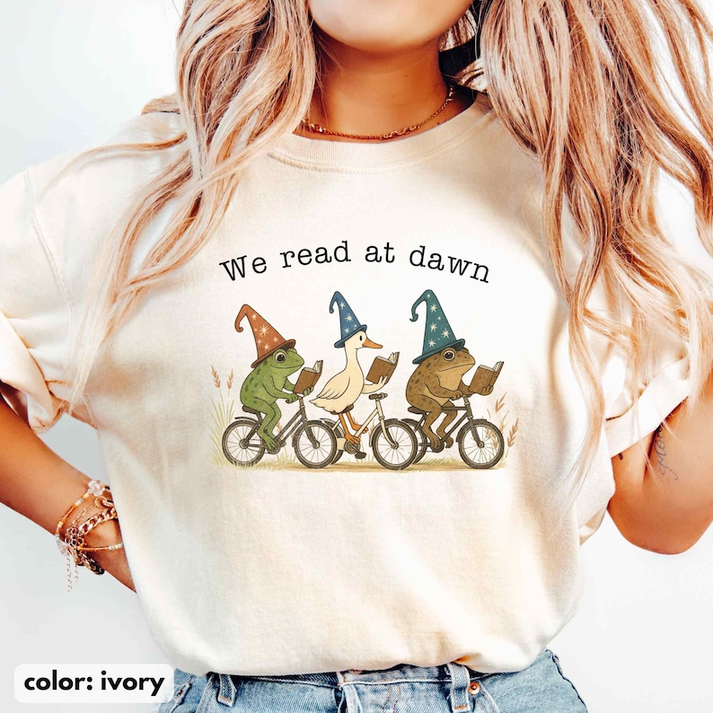 May include: Ivory-colored sweatshirt with the text "We read at dawn." The graphic features a duck and two frogs, all wearing wizard hats and riding bicycles while reading books. The color of the sweatshirt is ivory.