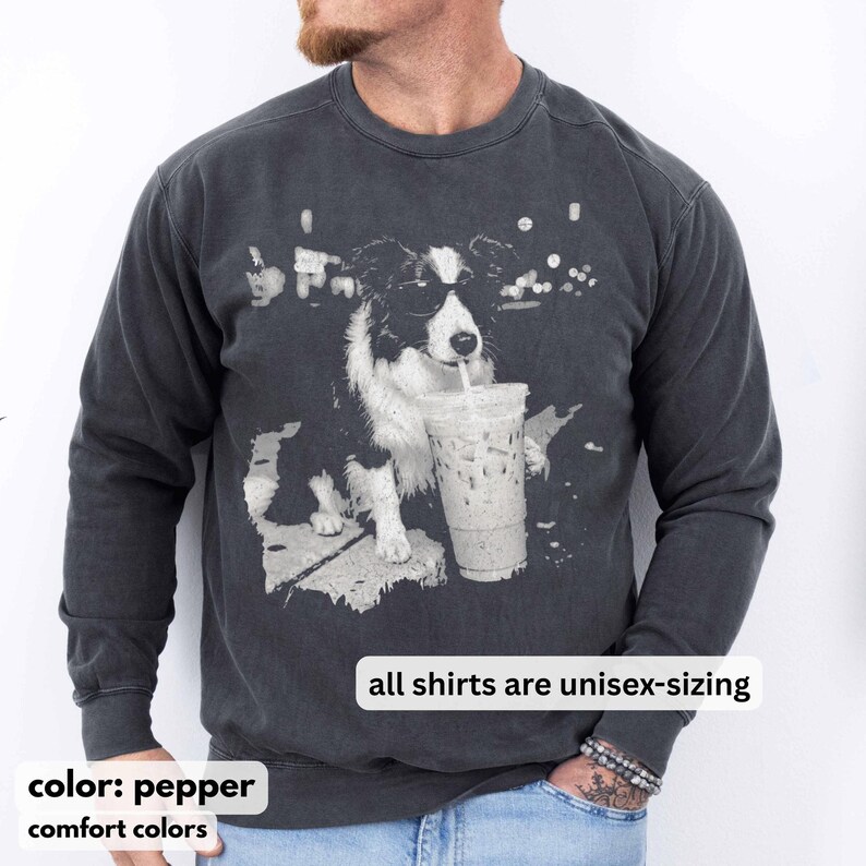 May include: Pepper-colored unisex sweatshirt featuring a black and white graphic of a dog drinking from a clear iced coffee cup. The text "all shirts are unisex-sizing" is displayed. The sweatshirt is part of the comfort colors collection.