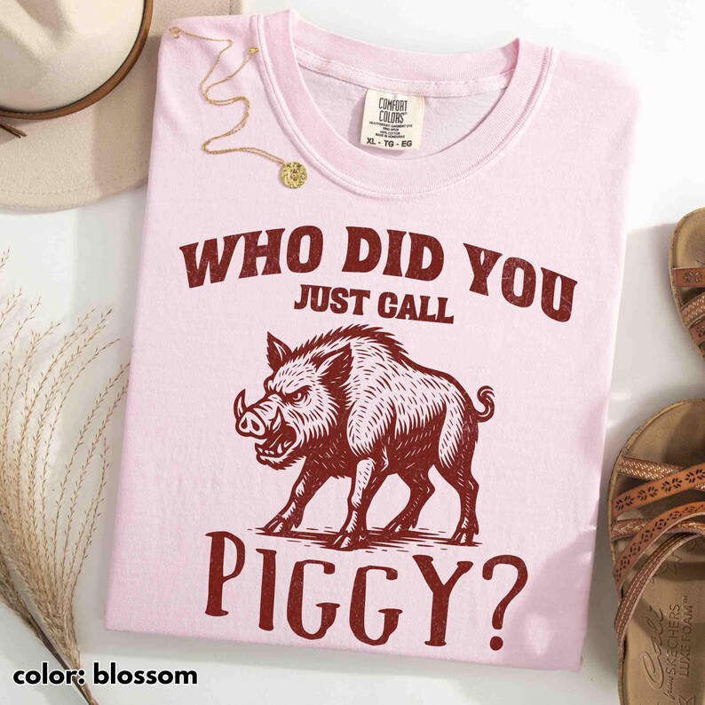 Op de afbeelding: Lichtroze t-shirt met de tekst "WHO DID YOU JUST CALL PIGGY?" en een donkerrode afbeelding van een wild zwijn. Het shirt is maat XL-TG-EG van Comfort Colors.
