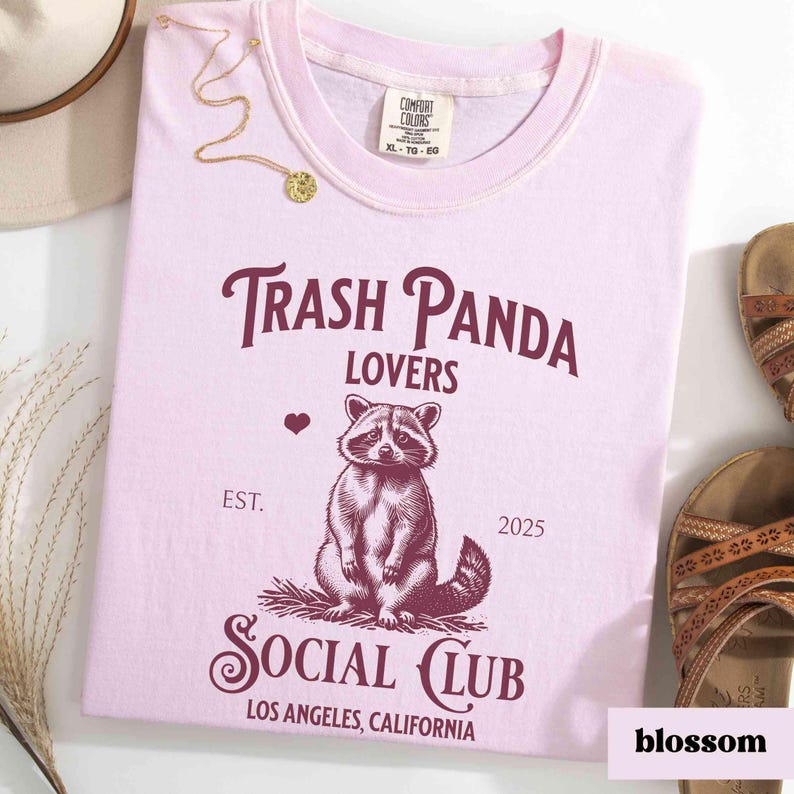 May include: Pink t-shirt with a graphic of a raccoon and the text "Trash Panda Lovers Social Club Est. 2025 Los Angeles, California"