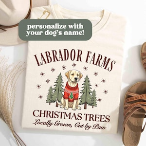 K&ouml;nnte beinhalten: Ein cremefarbenes T-Shirt mit einem weihnachtlichen Design, das einen Labrador Retriever zeigt, der einen roten Pullover mit einem kleinen Weihnachtsbaum tr&auml;gt. Der Text "Labrador Farms Christmas Trees Locally Grown, Cut by Paw" ist auf dem Shirt aufgedruckt.