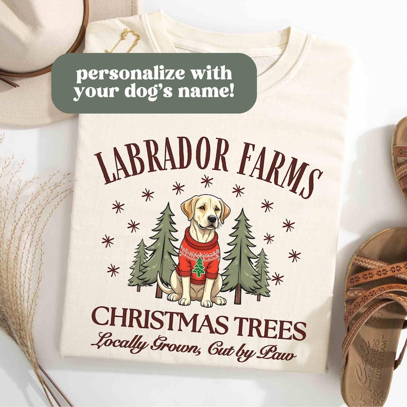 K&ouml;nnte beinhalten: Ein cremefarbenes T-Shirt mit einem weihnachtlichen Design, das einen Labrador Retriever zeigt, der einen roten Pullover mit einem kleinen Weihnachtsbaum tr&auml;gt. Der Text "Labrador Farms Christmas Trees Locally Grown, Cut by Paw" ist auf dem Shirt aufgedruckt.