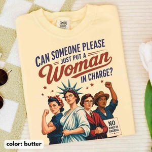 May include: Butter-colored t-shirt with the text "CAN SOMEONE PLEASE JUST PUT A WOMAN IN CHARGE?" and illustrations of women. The shirt also has the text "NO KINGS IN AMERICA."