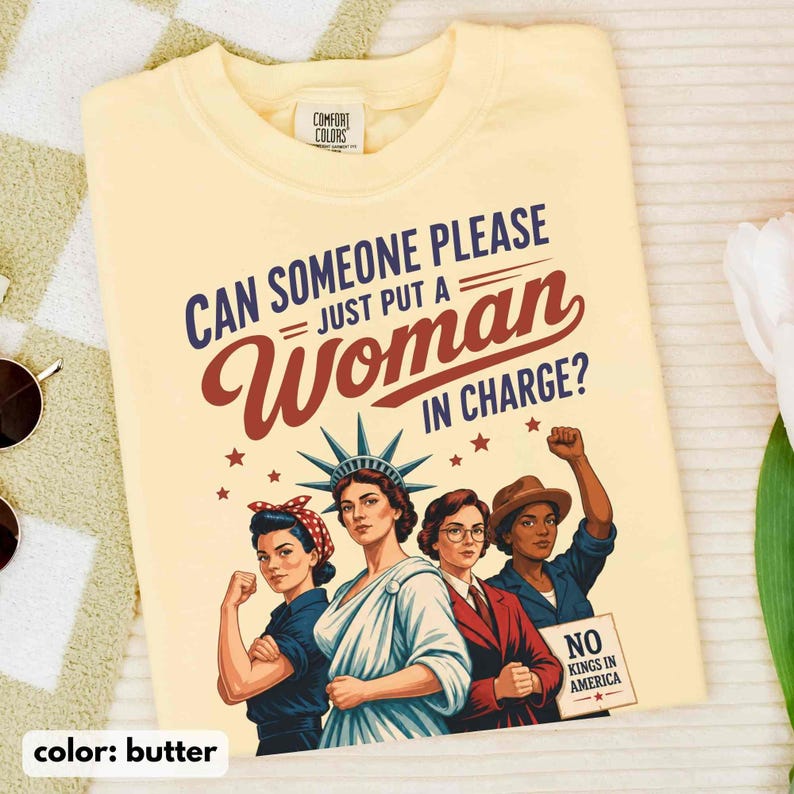 May include: Butter-colored t-shirt with the text "CAN SOMEONE PLEASE JUST PUT A WOMAN IN CHARGE?" and illustrations of women. The shirt also has the text "NO KINGS IN AMERICA."