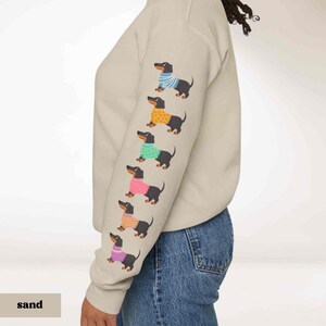 May include: A sand colored sweatshirt with a row of black dachshunds wearing colorful sweaters printed on the sleeve. The dachshund sweaters are blue, orange, green, pink, and purple.