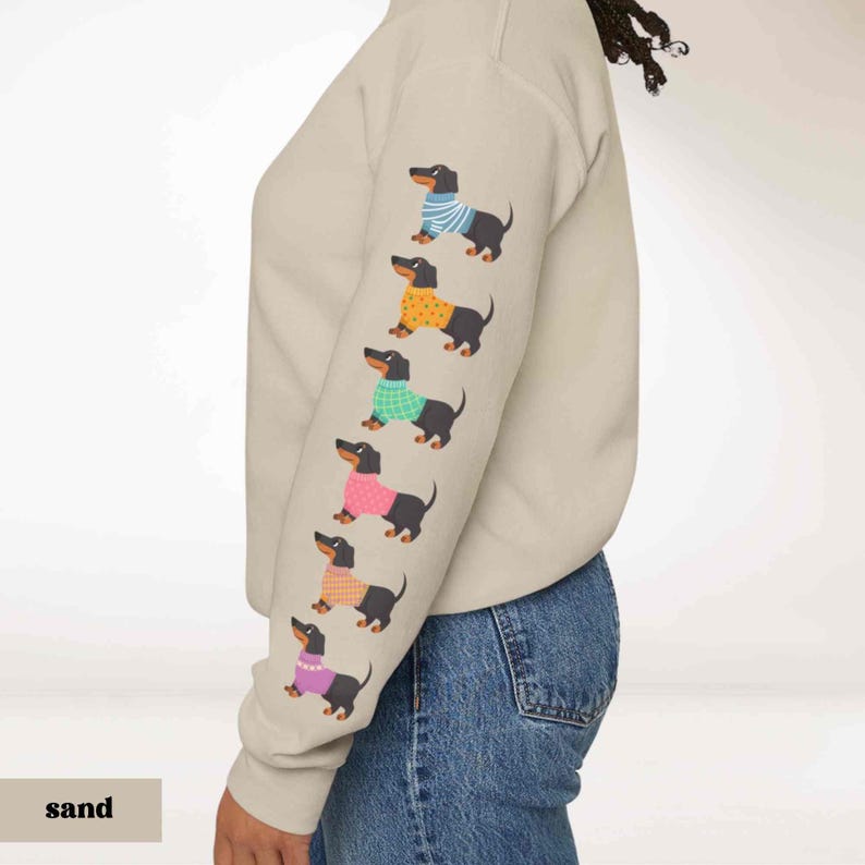 May include: A sand colored sweatshirt with a row of black dachshunds wearing colorful sweaters printed on the sleeve. The dachshund sweaters are blue, orange, green, pink, and purple.