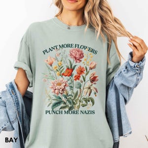 Op de afbeelding: Lichtgroen T-shirt met een bloemenontwerp met roze, rode en gele bloemen. De tekst "PLANT MORE FLOWERS" staat boven de bloemen en "PUNCH MORE NAZIS" eronder. Het shirt is gemaakt van een zacht materiaal.